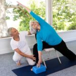 An older woman practicing a yoga pose using a chair and yoga block for support, guided by an instructor in a bright, spacious room with large windows overlooking trees.