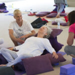 3 women in a yoga classroom studying a fourth woman in a reclined restorative yoga pose